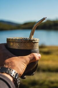 A hand holding a traditional yerba mate cup with a metal straw against a serene outdoor backdrop.