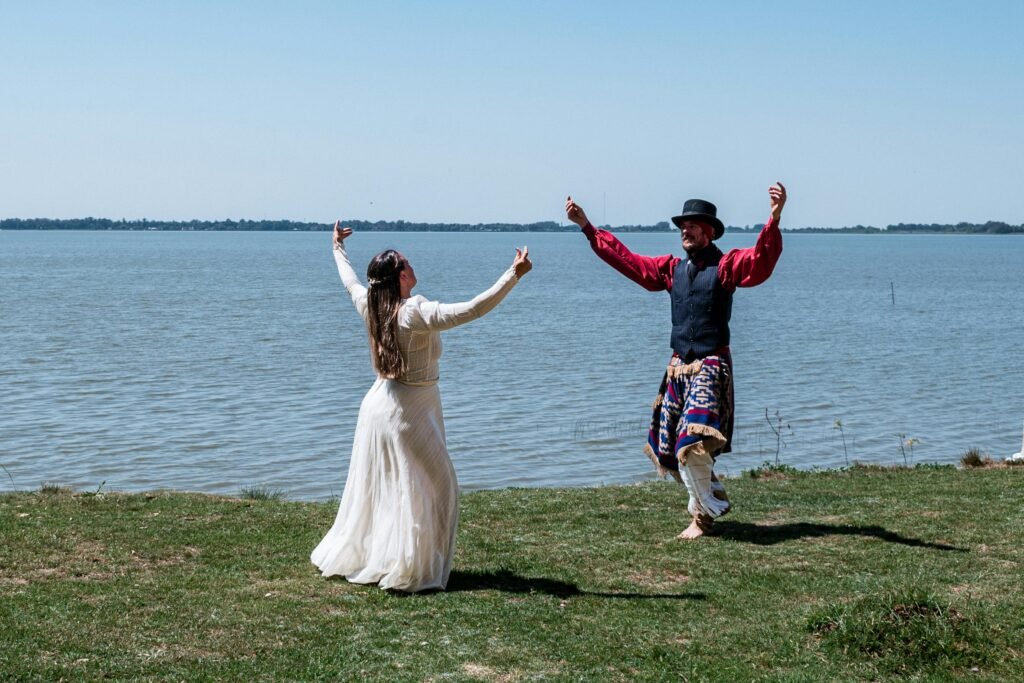 pexels-photo-29145607-29145607 Two people in traditional clothing dance by a lake in Chascomús, Argentina, under a clear blue sky.