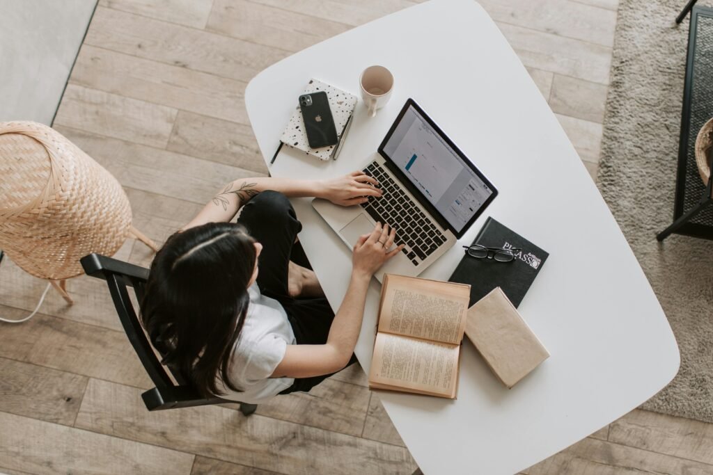 pexels-photo-4050315-4050315 Overhead view of a woman using a laptop at a home desk, surrounded by books, a phone, and a cup.